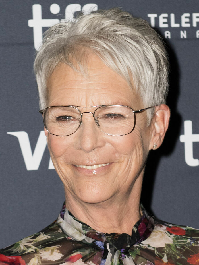 Jamie Lee Curtis smiling with short gray hair and glasses, wearing a floral top at a public event.