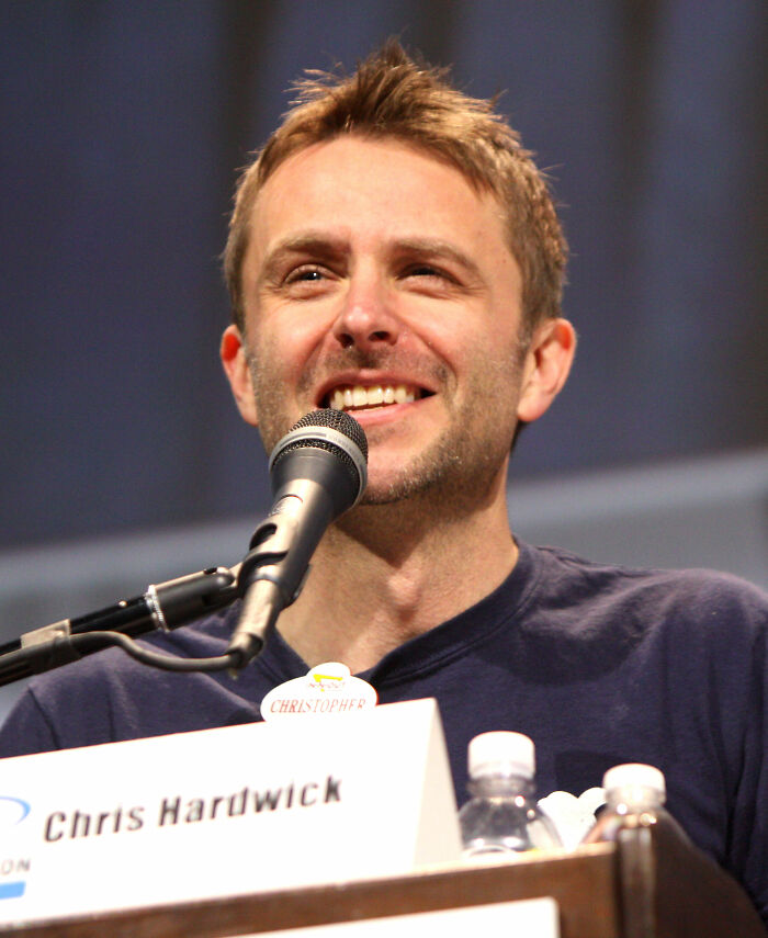 Chris Hardwick smiling while speaking into a microphone at a panel event, with nameplate and water bottles visible.