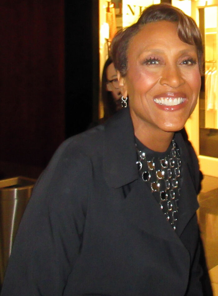 Robin Roberts smiling brightly, wearing a black coat and statement necklace, showcasing her career highlights.
