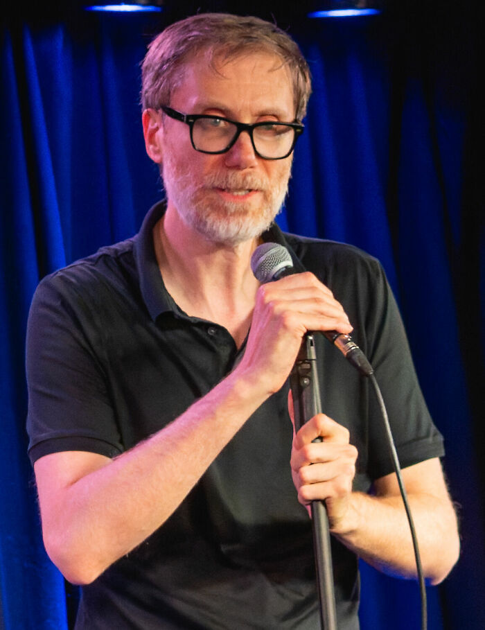 Stephen Merchant performing stand-up comedy on stage, holding a microphone with blue curtains in the background