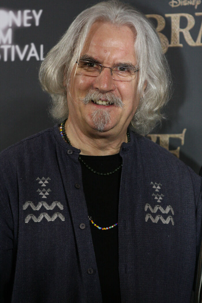 Billy Connolly smiling at an event, wearing glasses and a dark shirt with decorative patterns and beaded necklaces.