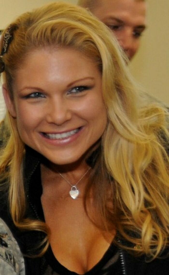 Beth Phoenix smiling with long blonde hair, wearing a black top and a silver pendant necklace in a casual setting