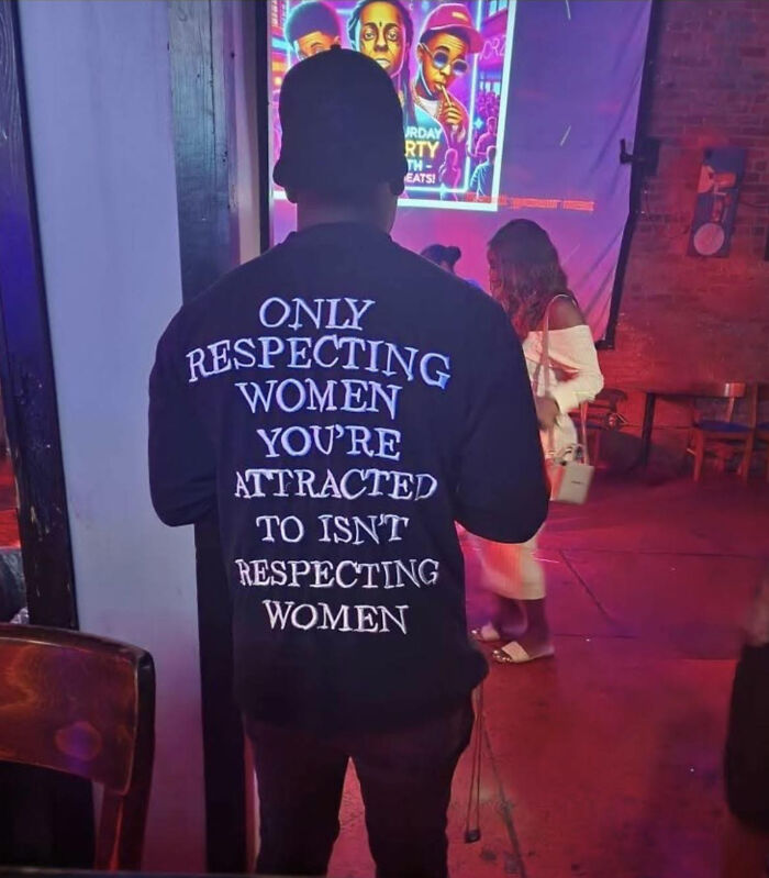Man wearing black shirt with text about respecting women, standing in dimly lit venue with people and colorful poster in background.