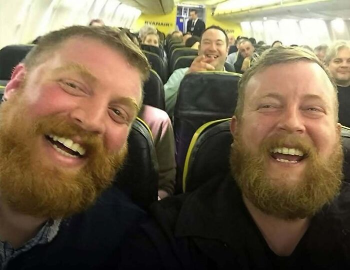 Two men with red beards smiling on a crowded airplane, showcasing candid moments from Spill Now photos and facts.