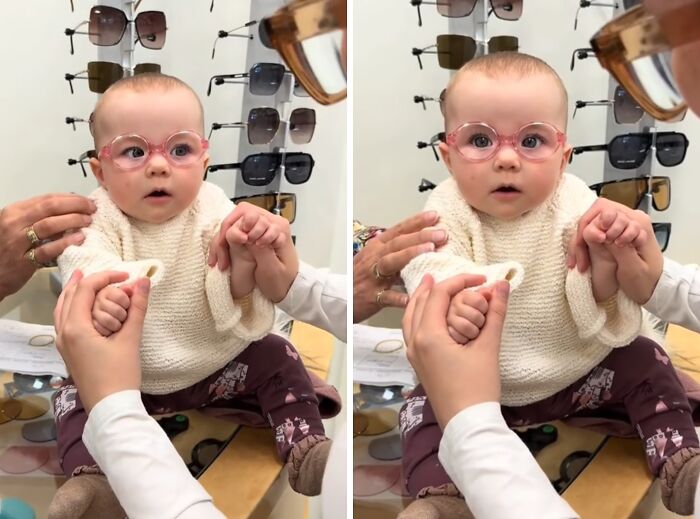 Baby wearing glasses sitting in front of a display of sunglasses, being supported by adult hands indoors in an optical shop