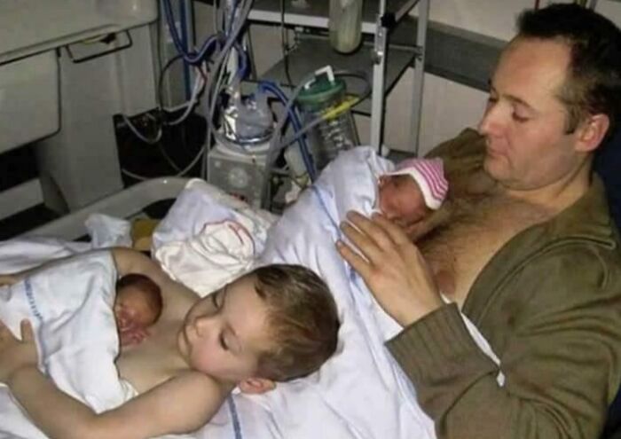 Father holding newborn twins while older child lies next to him, showing grateful sibling bond in hospital setting.