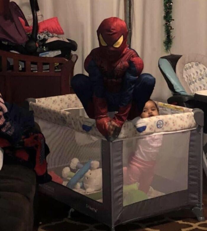Child dressed as Spider-Man sitting on a playpen edge while younger sibling stands inside smiling, showing sibling gratitude.