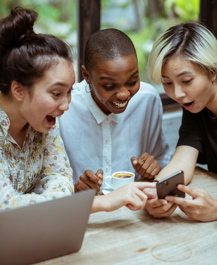 Three friends excitedly looking at a phone together, sharing moments of brutal revenge on an ex with zero regrets.