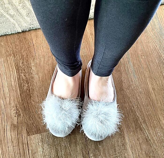Feet wearing black leggings and cozy slippers with fluffy pompoms, a pretty and practical aesthetic gift idea.