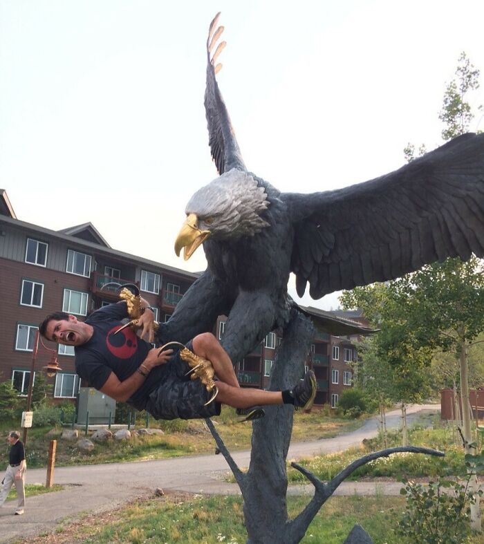 Hombre divirtiéndose al fingir ser atrapado por una escultura de águila, momento divertido de hombres que nunca crecieron.