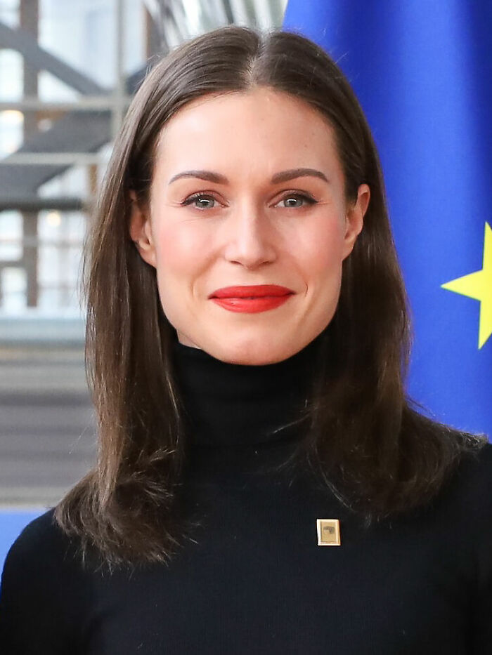 Sanna Marin wearing a black turtleneck and red lipstick, smiling in front of a blue European Union flag.