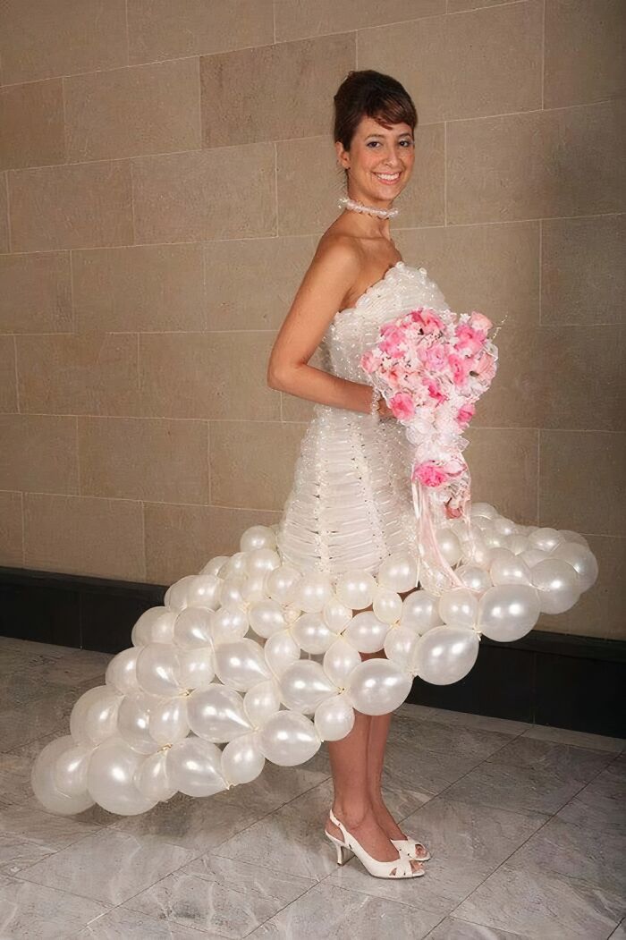 Bride in a unique wedding dress made of white balloons holding a pink floral bouquet, revealing questionable taste in fashion.
