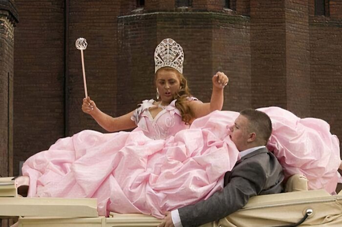 Bride in an exaggerated pink wedding dress with a large crown and wand, sitting next to a groom in a vintage car.