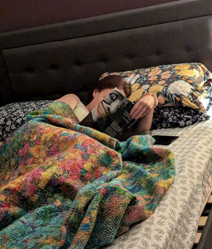 Person lying in bed under a colorful quilt, holding a book with a cover that perfectly aligns with their face.