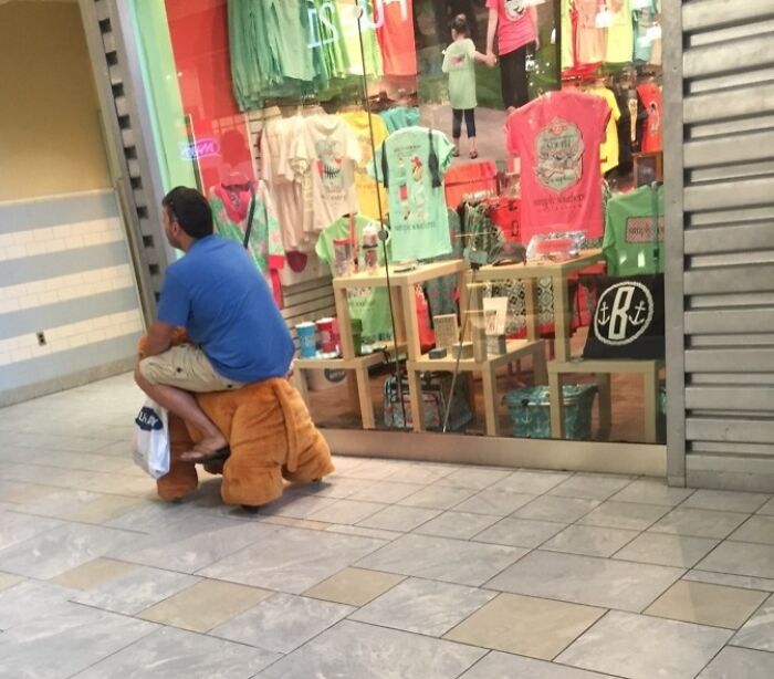 Man wearing blue shirt sitting on a plush toy ride-on in a mall, showing wholesome men never grow up behavior.