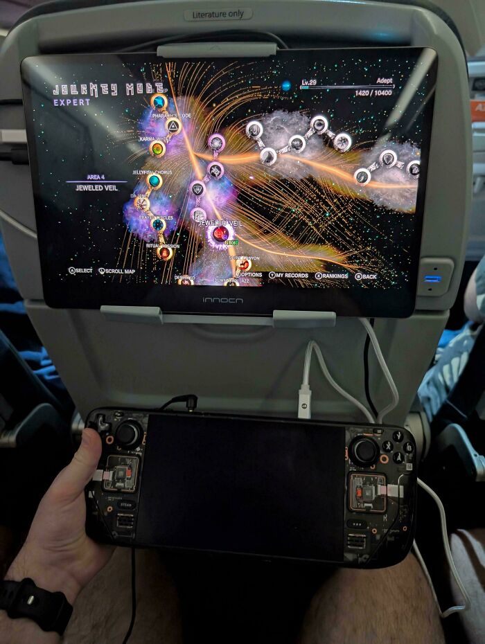 Hand holding a portable gaming console aligned perfectly with an in-flight entertainment screen showing a game map.