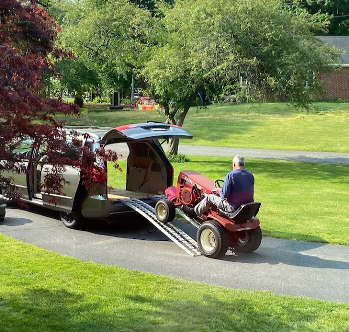 Man driving riding lawn mower onto van ramp in a green yard, showing men never grow up in wholesome moment.