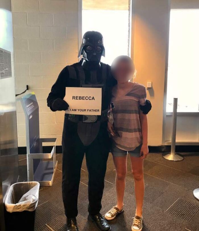 Hombre disfrazado de Darth Vader con cartel humorístico posando con mujer mostrando momentos divertidos de hombres que nunca crecieron.