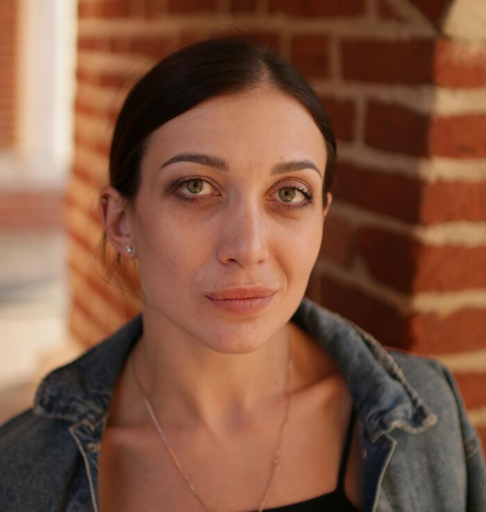 Young woman with tired eyes and casual denim jacket leaning against brick wall, showing relatable emotions and expressions.