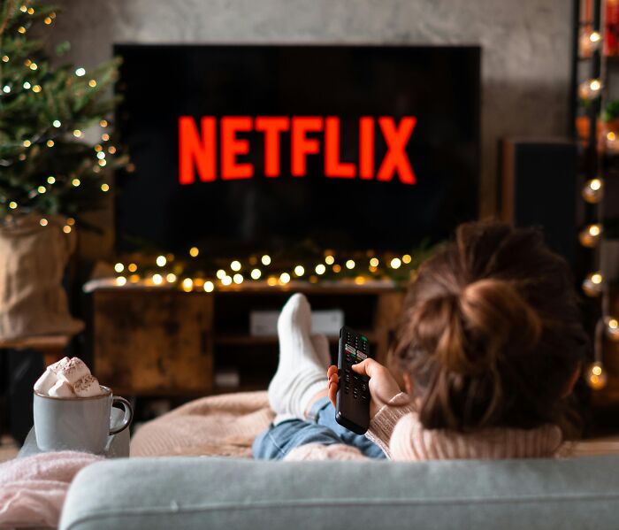 Person relaxing at home watching Netflix with remote in hand and hot chocolate nearby, relatable guilty pleasures.