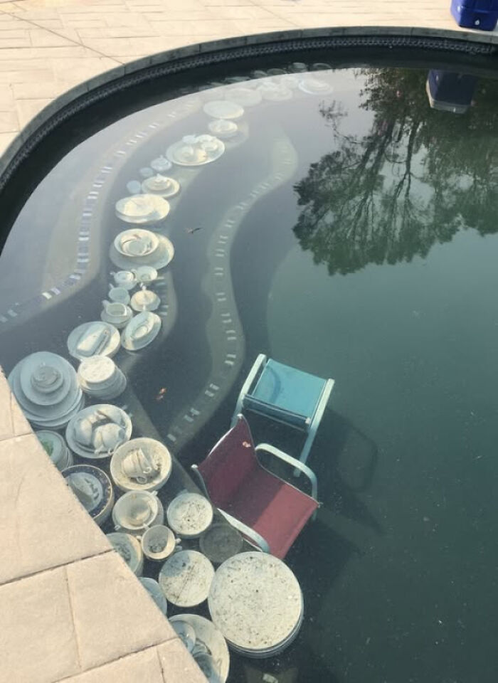 Partially submerged chairs and white dishware in a pool reflecting trees, evoking existence and reality themes.