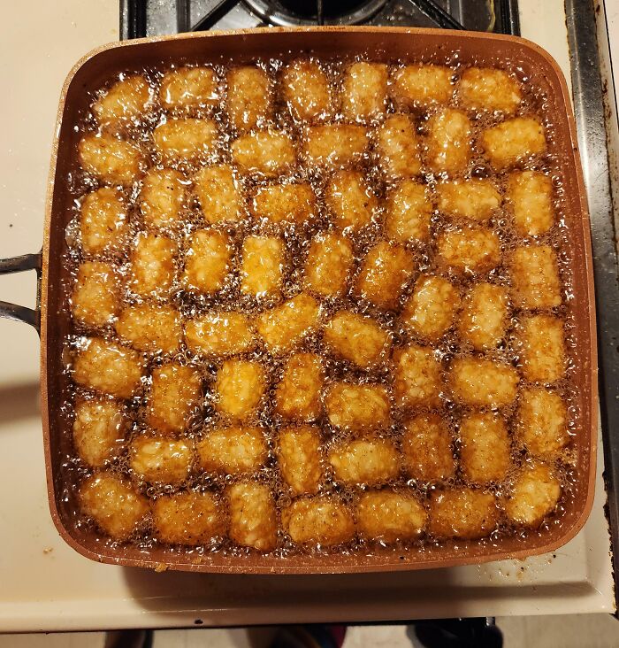 Frozen tater tots perfectly aligned in a square pan, frying evenly in hot oil on a stovetop burner.