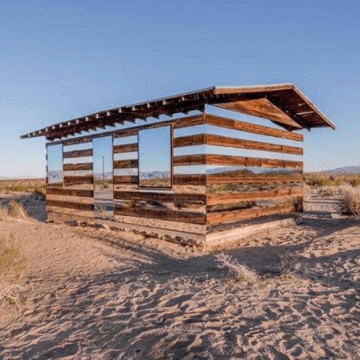A wooden house with mirrored walls reflecting the desert landscape, illustrating concepts of existence and reality.
