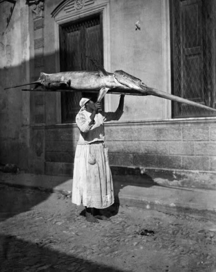 Woman carrying a large swordfish on her head in a street, illustrating existence and reality in the universe context.