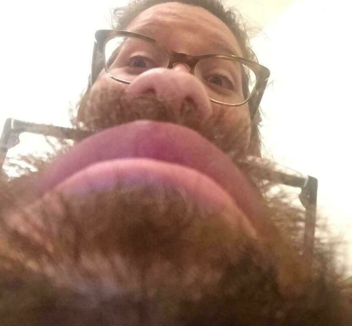 Close-up photo from below of a man wearing glasses with a beard that makes the image confusing to the brain.
