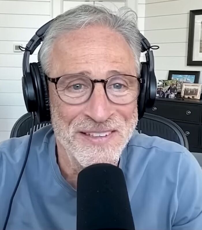 Jon Stewart with headphones and glasses speaking into a microphone during a podcast or interview session.