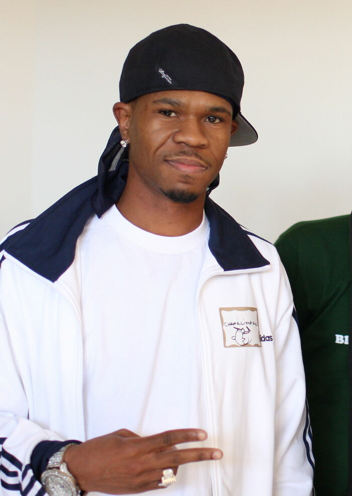 Chamillionaire wearing a black cap and white jacket, posing with a peace sign indoors.
