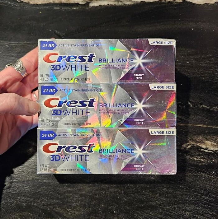 Hand holding three large-size Crest 3D White Brilliance toothpaste boxes, featured in top Cyber Monday deals.