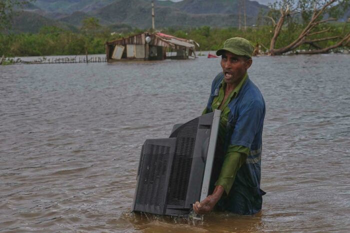 Man wading through floodwaters holding a large broken TV, showcasing strength and resilience in challenging conditions.