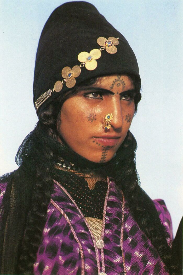 Portrait of a woman with traditional facial tattoos and jewelry wearing a black headpiece and purple garment showcasing human diversity.