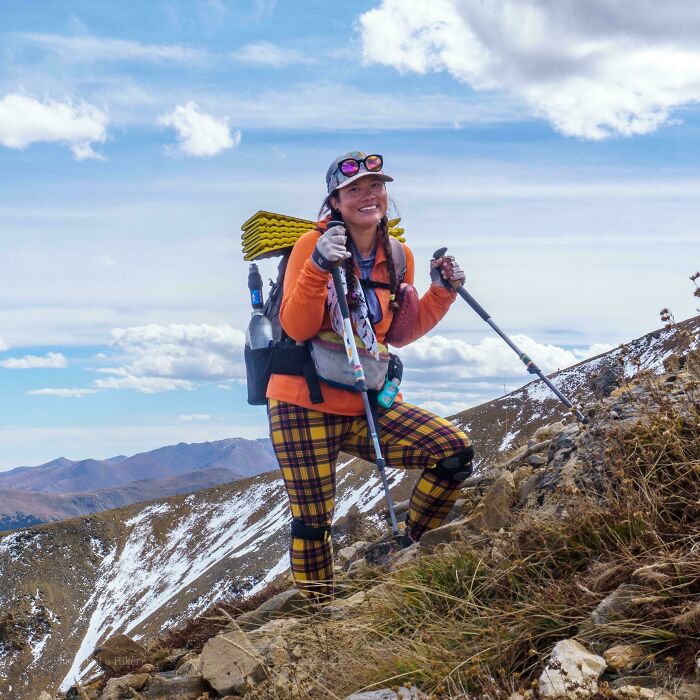 Hiker wearing orange and plaid, using trekking poles on a mountain trail, showcasing human strength and diversity.