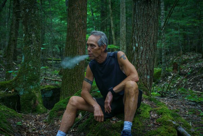 Middle-aged man with tattoos sitting in a forest, exhaling smoke, capturing the strength and diversity of humans.
