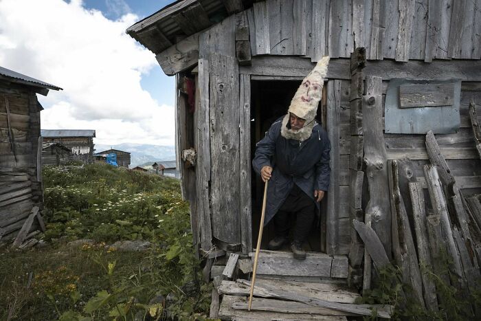 Elderly person wearing traditional tall hat and holding walking stick, exiting rustic wooden house in rural setting, capturing human diversity.