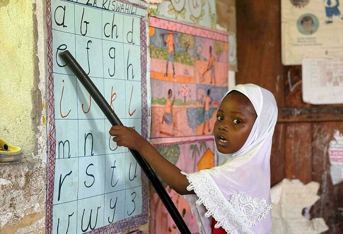 Young girl pointing to letters on a Kiswahili alphabet chart, showcasing the diversity and strength of humans in education.