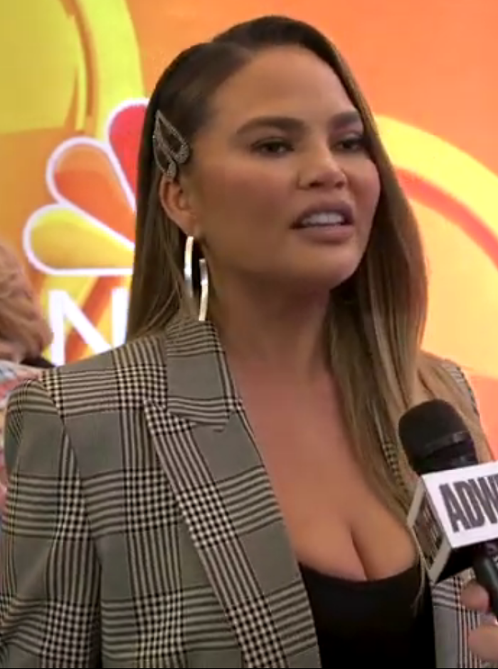 Chrissy Teigen speaking in an interview, wearing plaid blazer and hoop earrings at a media event background.