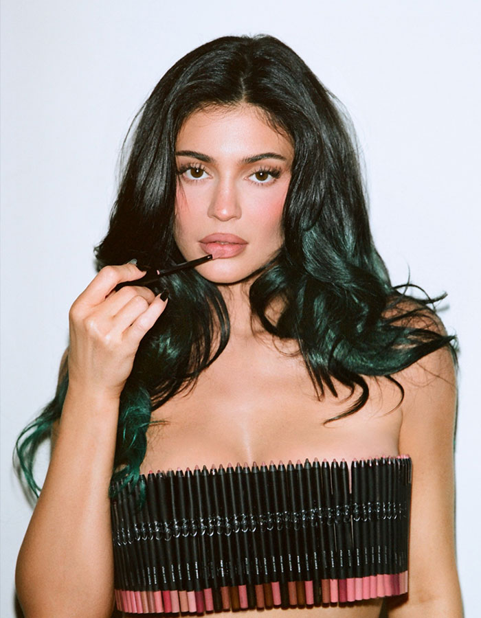 Young woman with long dark hair applying lip liner, reflecting Kris Jenner family photo debate on most annoying family member.