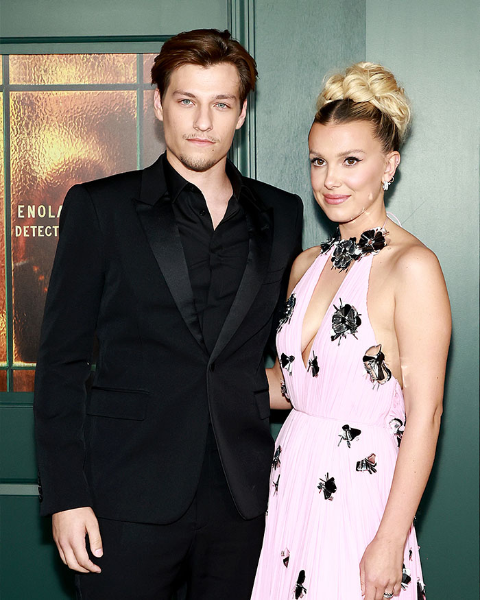 Millie Bobby Brown posing with a man at an event, dressed in a pink floral dress amid adoption and criticism discussions. Millie Bobby Brown posing with a man at an event, dressed in a pink floral dress amid adoption and criticism discussions.