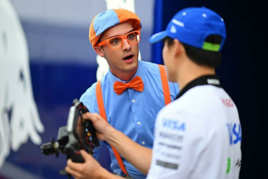 Actor dressed as Blippi wearing an orange bow tie and cap talking to a man holding a camera remote control.