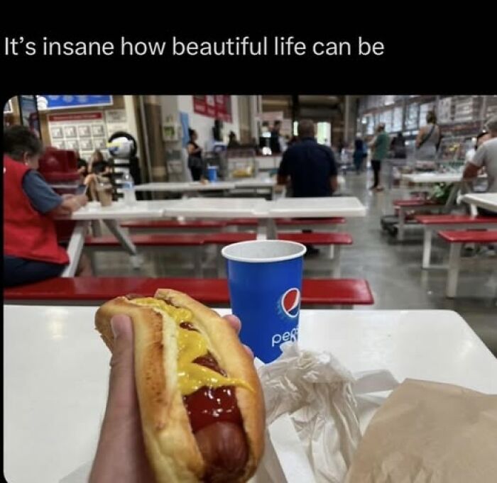 Hand holding a hot dog with mustard and ketchup, a Pepsi cup on the table, showcasing popular pics that go hard in a food court.