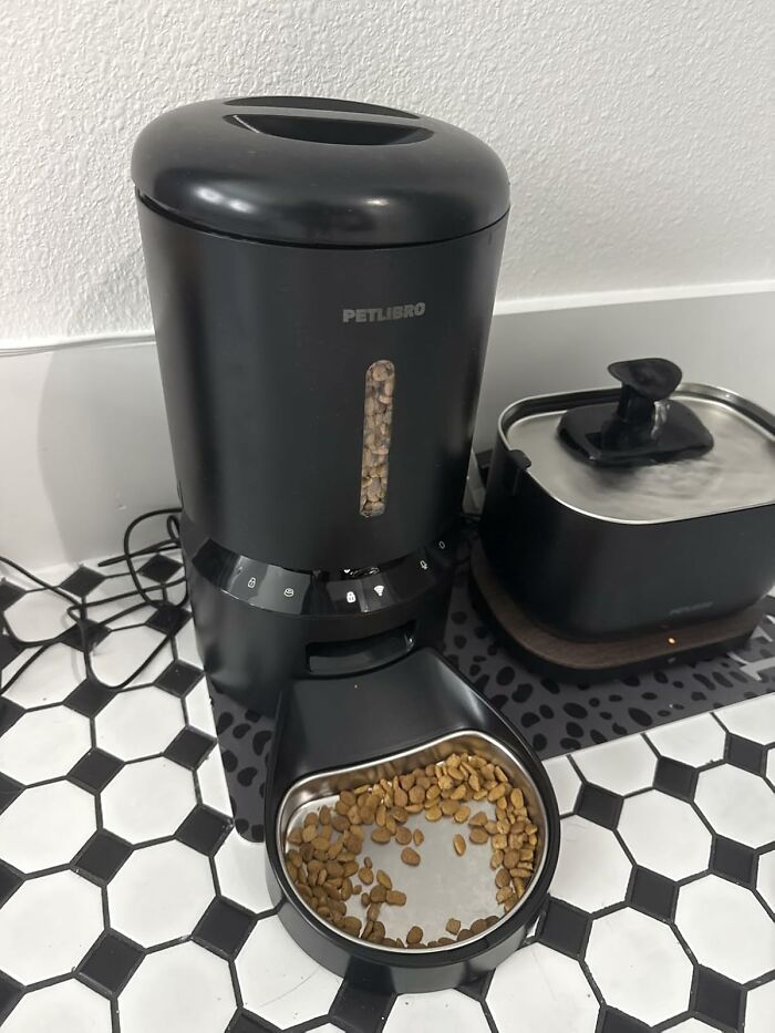 Black automatic pet feeder dispensing dry food, pet care product trending on TikTok, placed on patterned floor tiles.