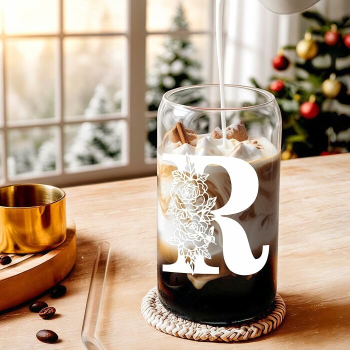 Cold coffee drink with cream and cinnamon in a floral R monogram glass, set on a wooden table near a window with winter decor.