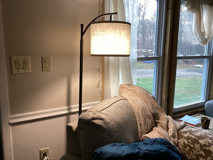 Cozy living room corner with glowing floor lamp, soft blanket on sofa, and natural light through large windows.