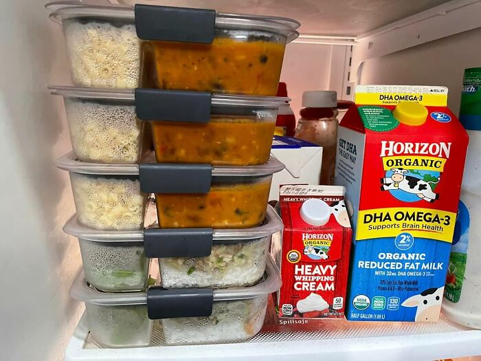 Stacked meal prep containers with soups and sides inside a fridge alongside organic milk and heavy whipping cream.