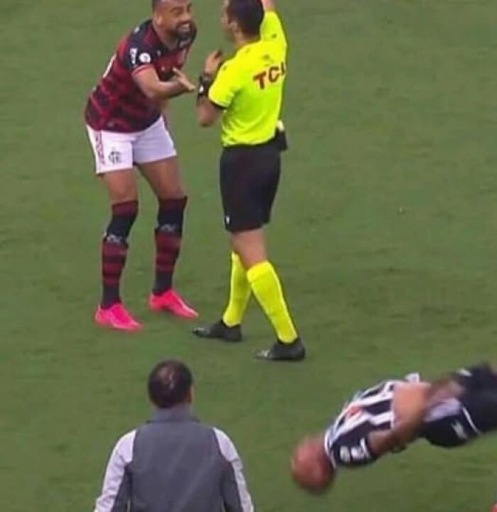 Soccer player reacting to referee on field while another player does a dramatic flip during a hard-hitting match moment.