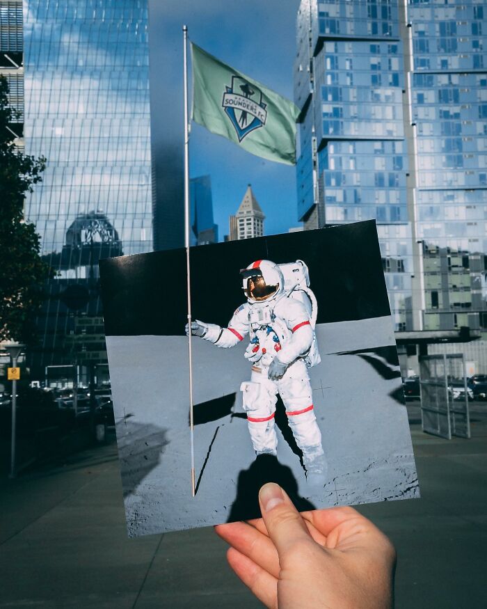 Hand holding a paper cutout of an astronaut interacting with a real flagpole in an urban cityscape.