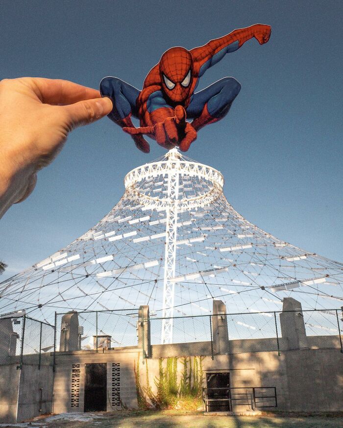 Hand holding paper cutout of Spider-Man appearing to interact with a white tower structure in daylight, paper cutouts.